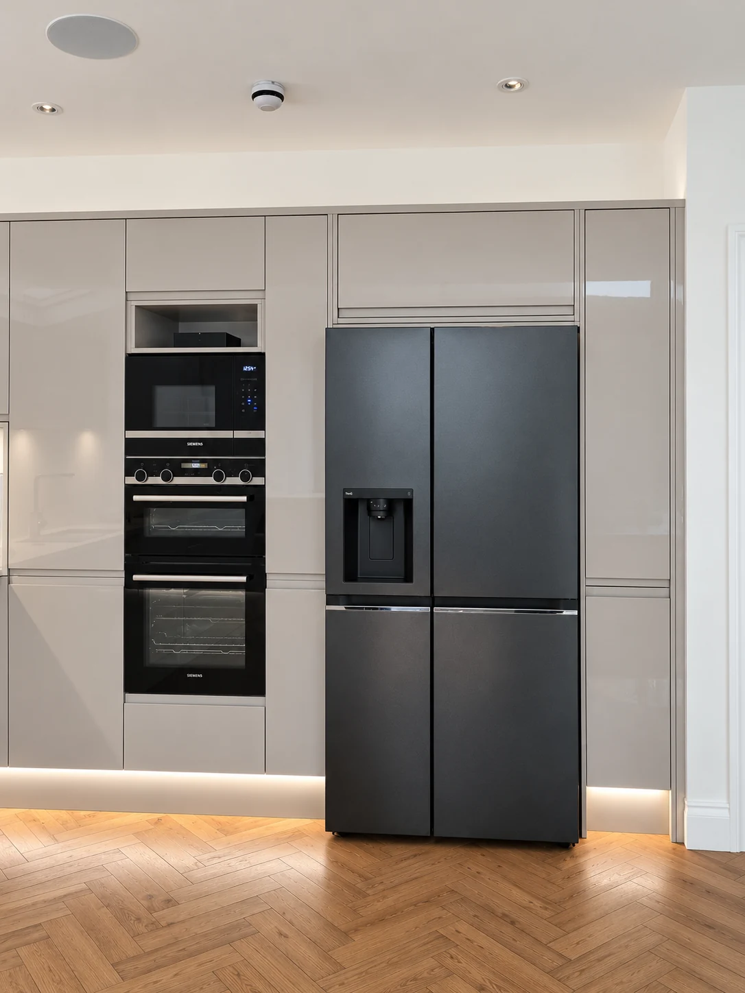 Tall grey kitchen cabinetry with stacked cooking appliances, dark refrigerator and a herringbone timber floor.