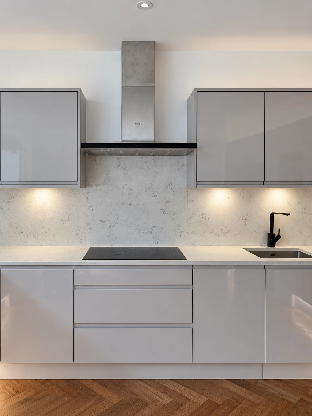 Kitchen hob wall with grey gloss cupboards, pale veined splashback and a sink to the right.