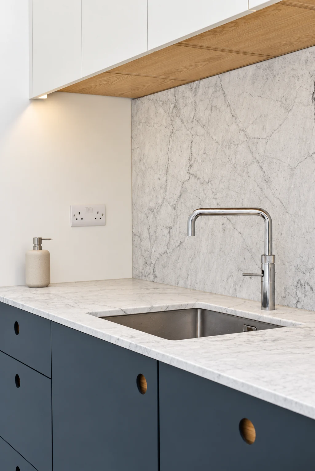 Veined stone worktop, stainless steel sink and chrome tap above dark blue cabinets with recessed finger pulls.