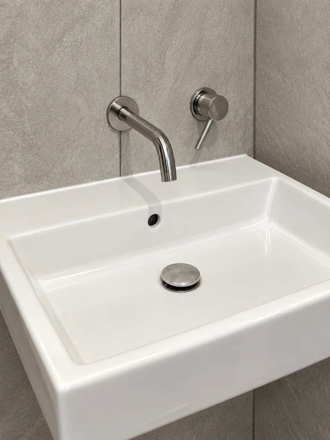 White rectangular basin beneath a wall-mounted spout and separate mixer, against large grey tiles.