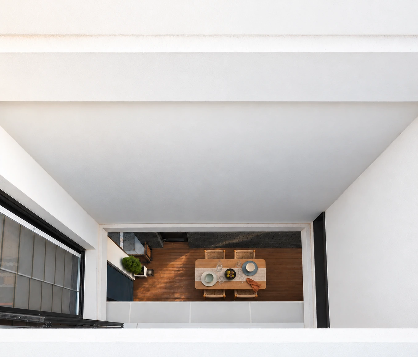 View down a white lightwell to a timber courtyard table with four woven chairs and a simple lunch setting.