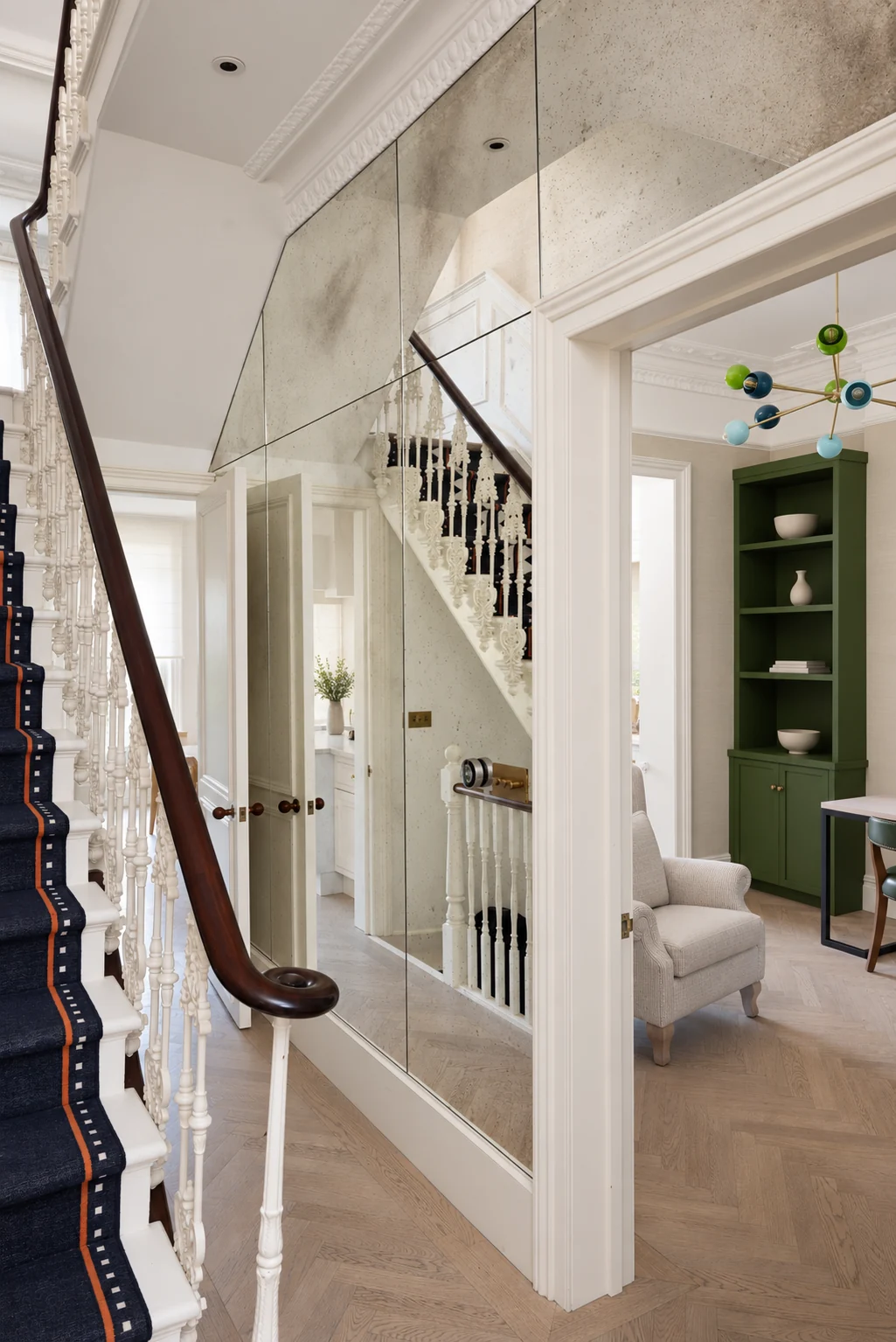 Curved antiqued mirror beside an ornate white stair, dark handrail and green library.